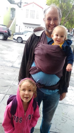 Hamilton with a baby in a wrap and a young kid in a pink hoodie on a San Francisco sidewalk